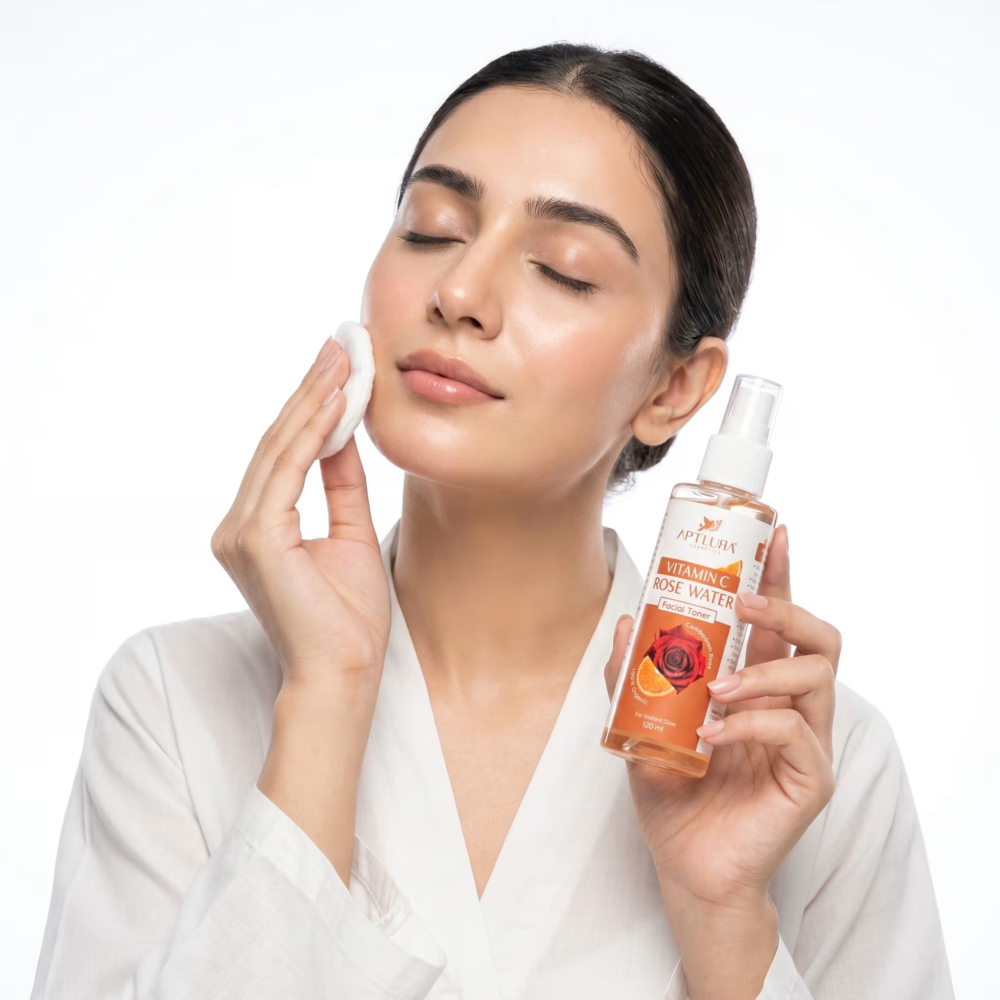 Woman applying rose water to her face with a bottle of rose water in her hand on a white background