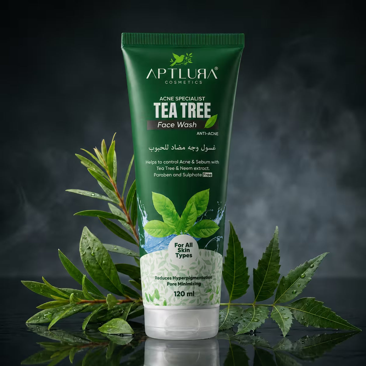 A 120 ml tube of Aptlura Cosmetics Tea Tree Face Wash stands upright on a reflective black surface, surrounded by fresh green tea tree leaves. The background is a dark, smoky studio setting.