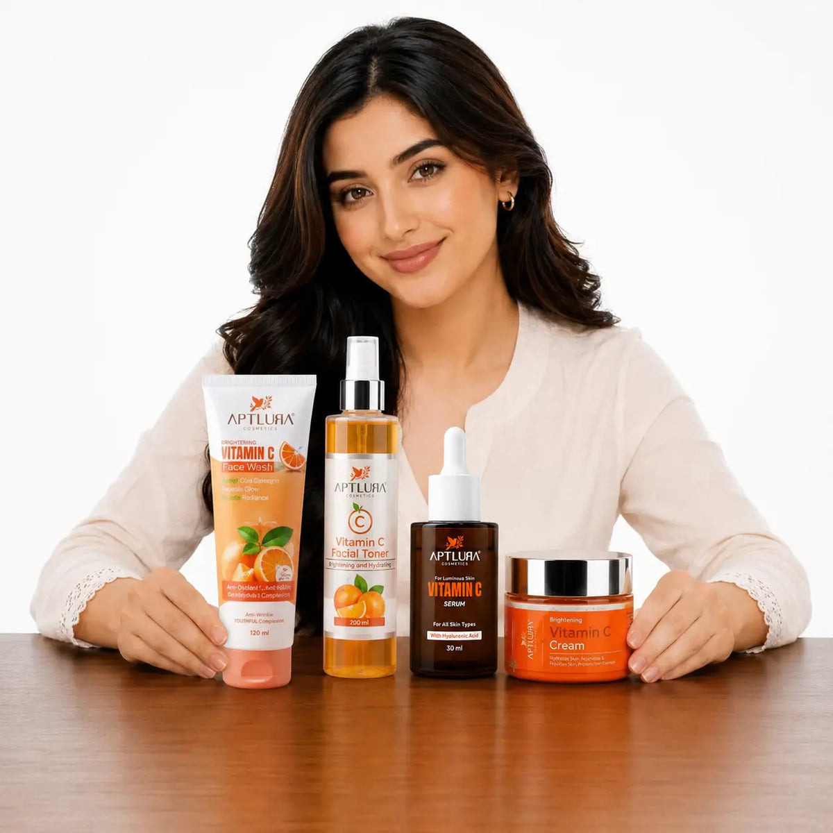 A woman seated at a wooden table with four Aptlura Cosmetics products in front of her on a white studio background. The products include a Vitamin C Hand Cream tube, a Vitamin C Serum bottle, a Vitamin C Cream dropper bottle, and a Vitamin C Cream jar.