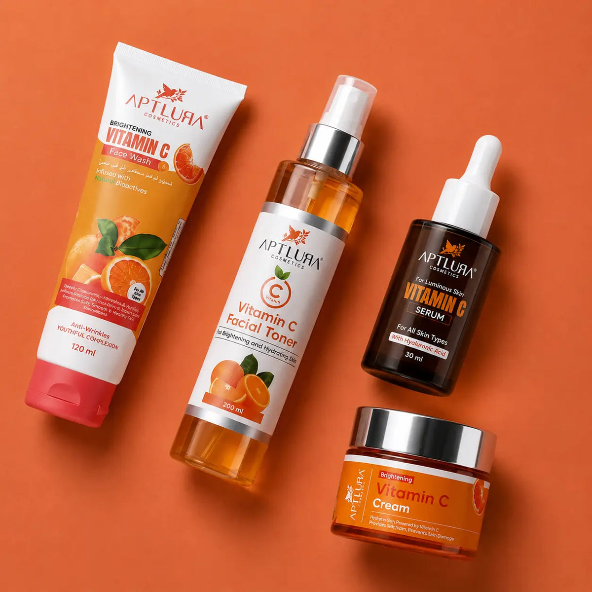 Four skincare products from Aptlura cosmetics arranged on a solid orange background: a Vitamin C Brightening Hand Cream tube, a Vitamin C Facial Toner bottle, a Vitamin C Serum dropper bottle, and a Vitamin C Cream jar.