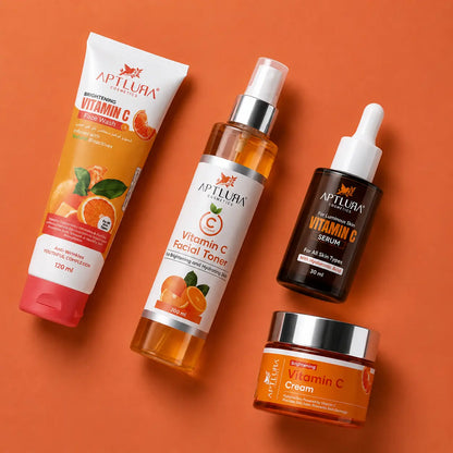 Four skincare products from Aptlura cosmetics arranged on a solid orange background: a Vitamin C Brightening Hand Cream tube, a Vitamin C Facial Toner bottle, a Vitamin C Serum dropper bottle, and a Vitamin C Cream jar.