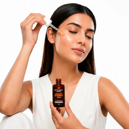 A woman with closed eyes applies a golden serum droplet from a dropper to her cheek while holding a brown bottle labeled 'APTLURA Vitamin C Serum' against a clean white background.